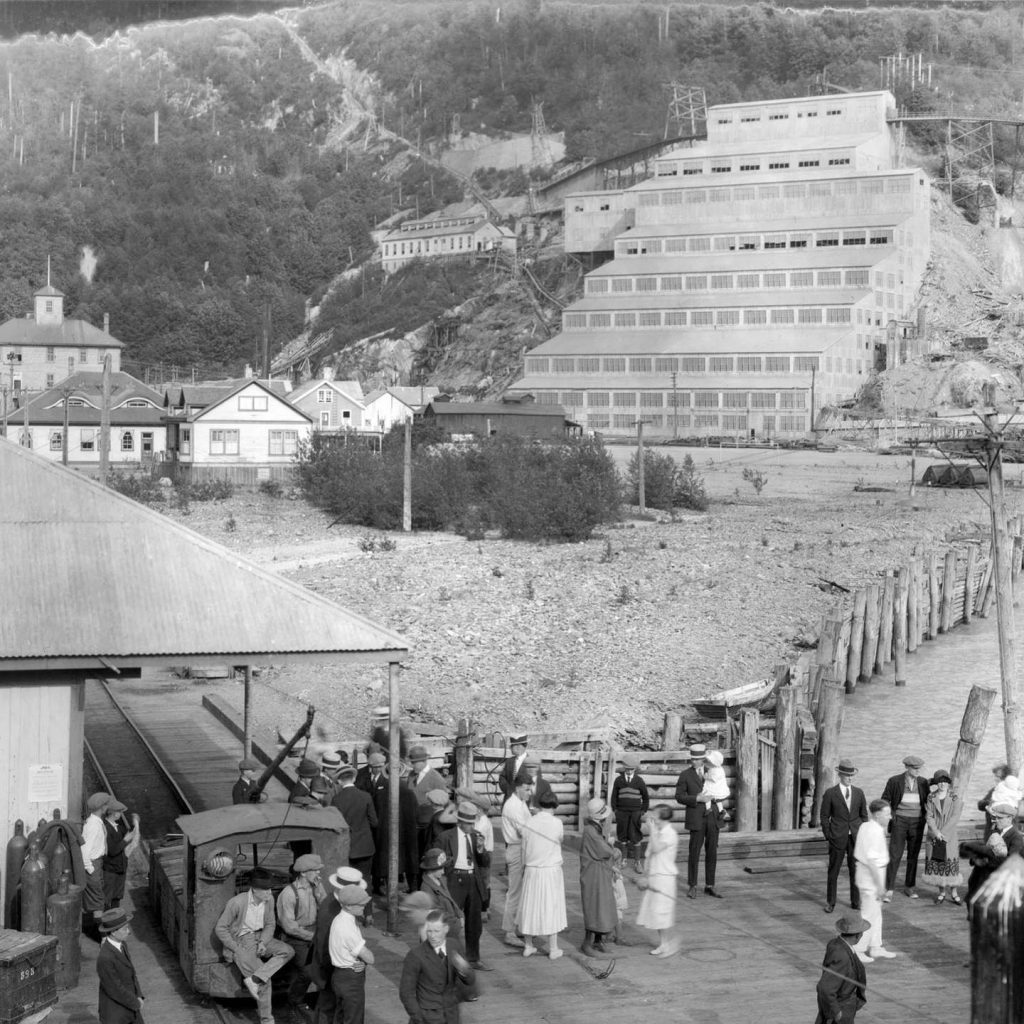 Black and white photo of men and women waiting for the steam train with Britannia Mine in the background in the early 1900’s